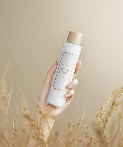 Hand holding a bottle of rice toner with a beige background and dry grass at the bottom.