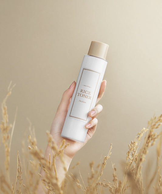 Hand holding a bottle of rice toner with a beige background and dry grass at the bottom.