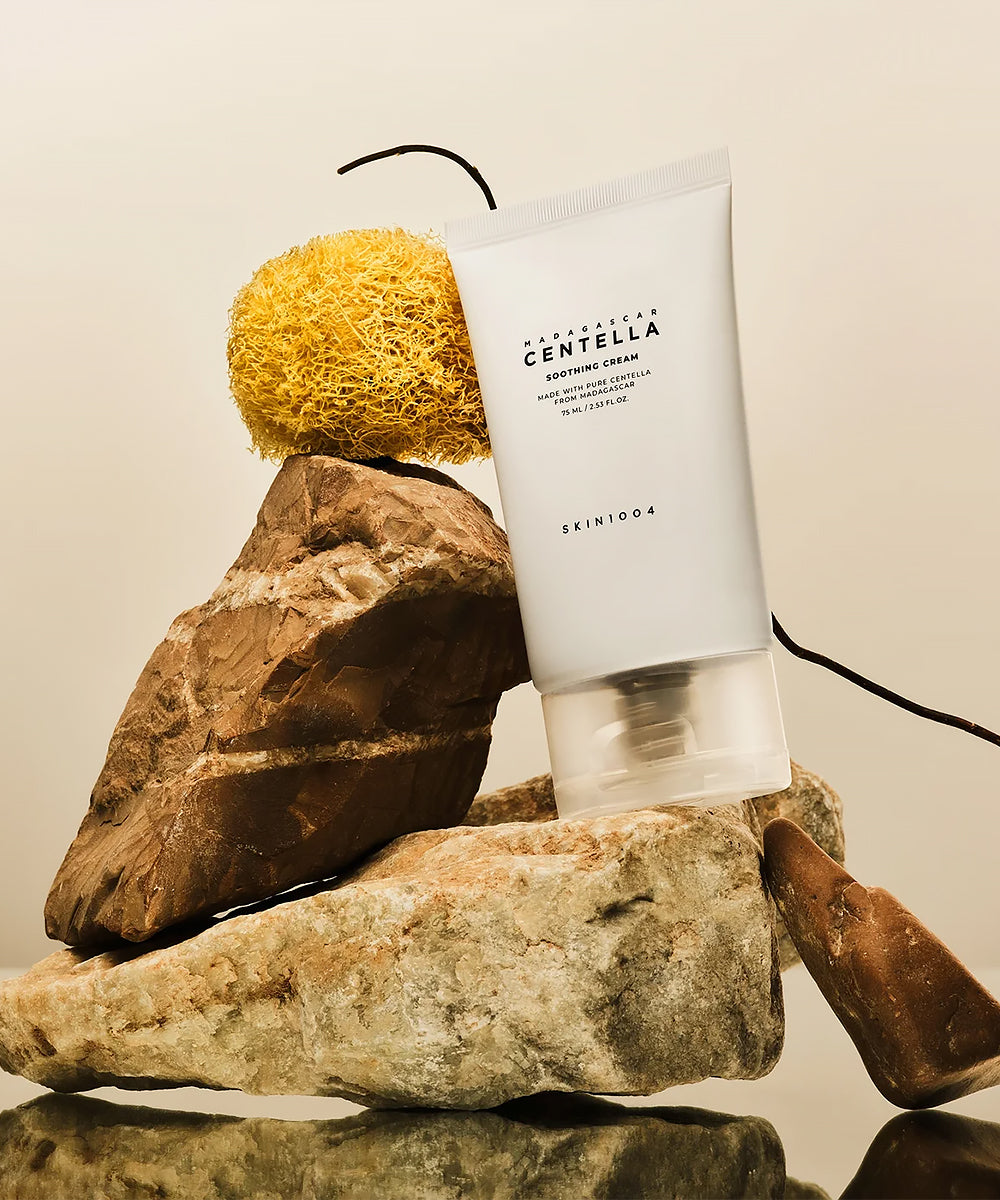 The SKIN1004 Madagascar Centella Soothing Cream tube aesthetically displayed on a stack of rough natural rocks with a natural sponge and dried vine