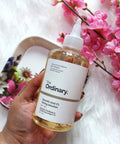 Hand holding a bottle of 'The Ordinary' Glycolic Acid 7% Toning Solution with pink flowers in the background.