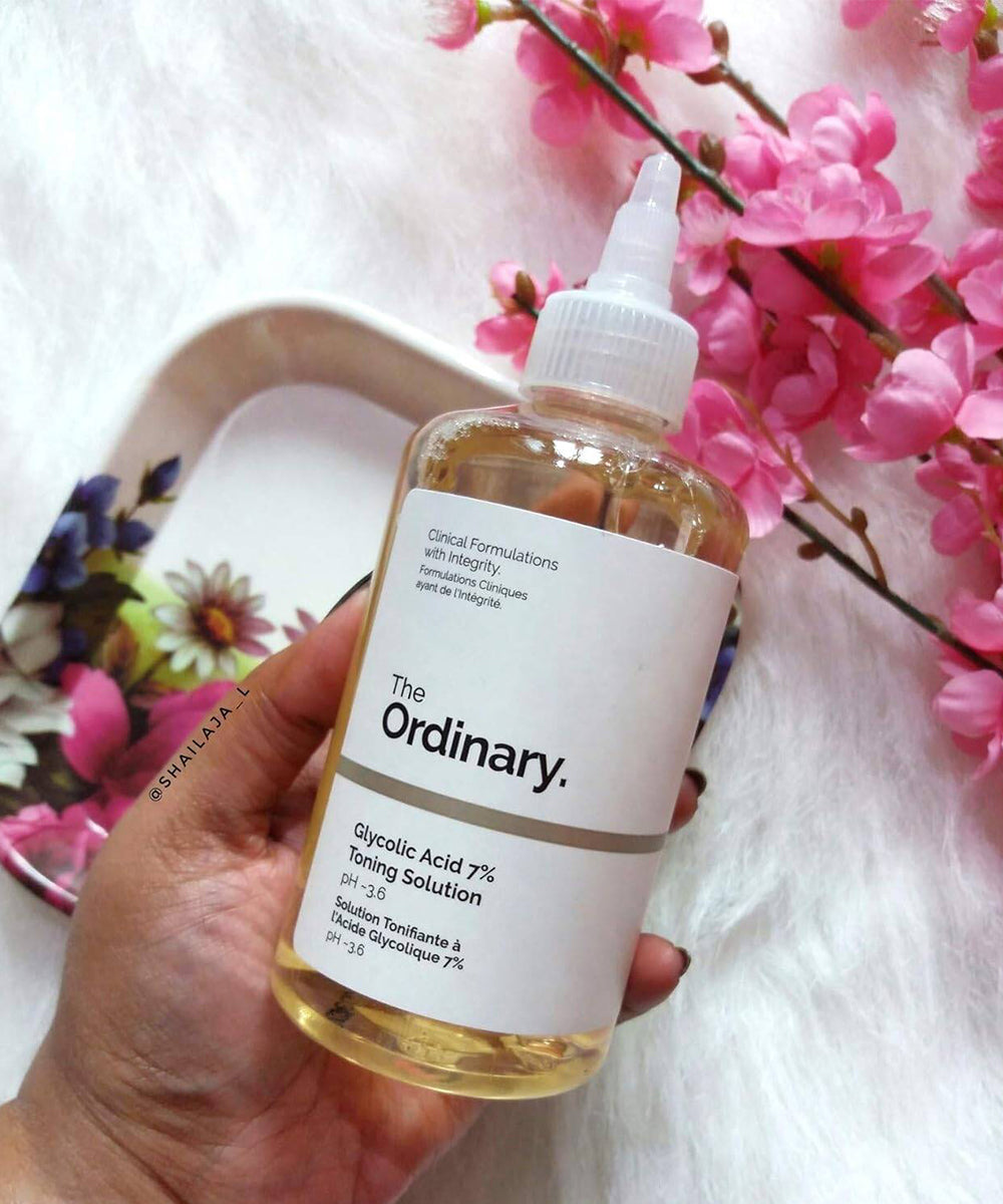 Hand holding a bottle of 'The Ordinary' Glycolic Acid 7% Toning Solution with pink flowers in the background.