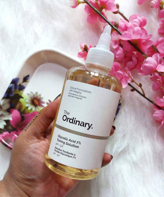 Hand holding a bottle of 'The Ordinary' Glycolic Acid 7% Toning Solution with pink flowers in the background.