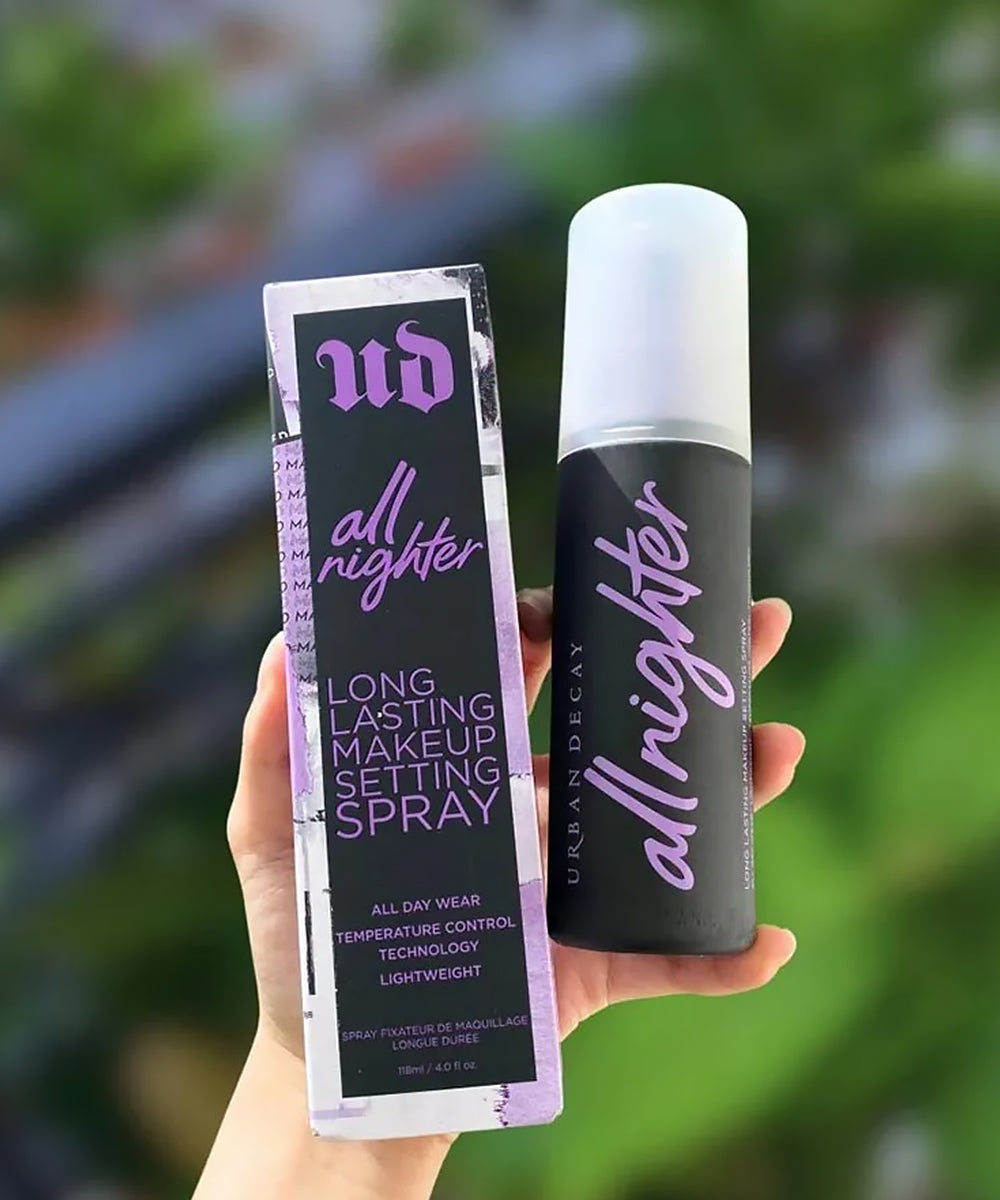 Hand holding Urban Decay 'all nighter' makeup setting spray against a blurred green background