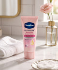 Vaseline Glutaglow Ampoule Ultra Bright dry cream dehydrating treatment tube, set on a marble countertop with a white towel, a gold-rimmed mirror, and a pink rose in a vase in the background.